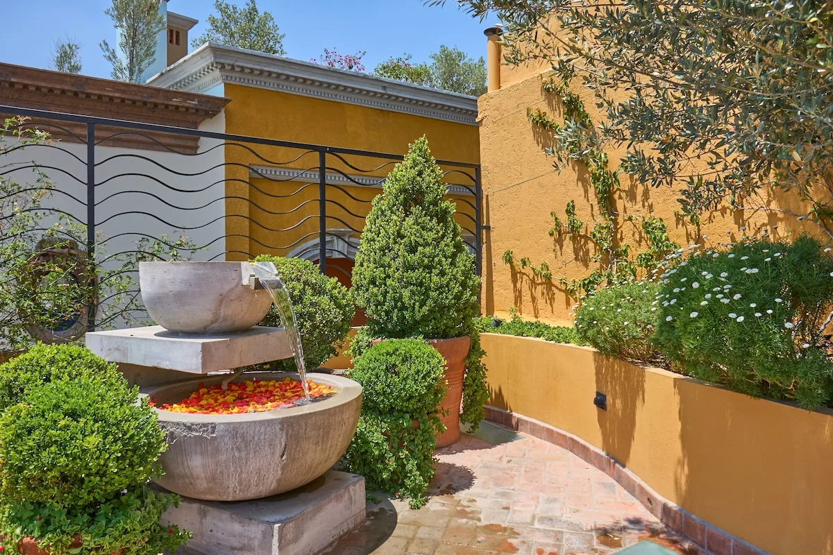 Un jardín de patio presenta una fuente de piedra, arbustos podados y muros de tonos cálidos.