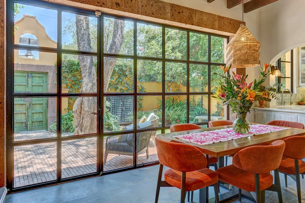 Un comedor con una mesa de madera y asientos coloridos junto a grandes ventanas.