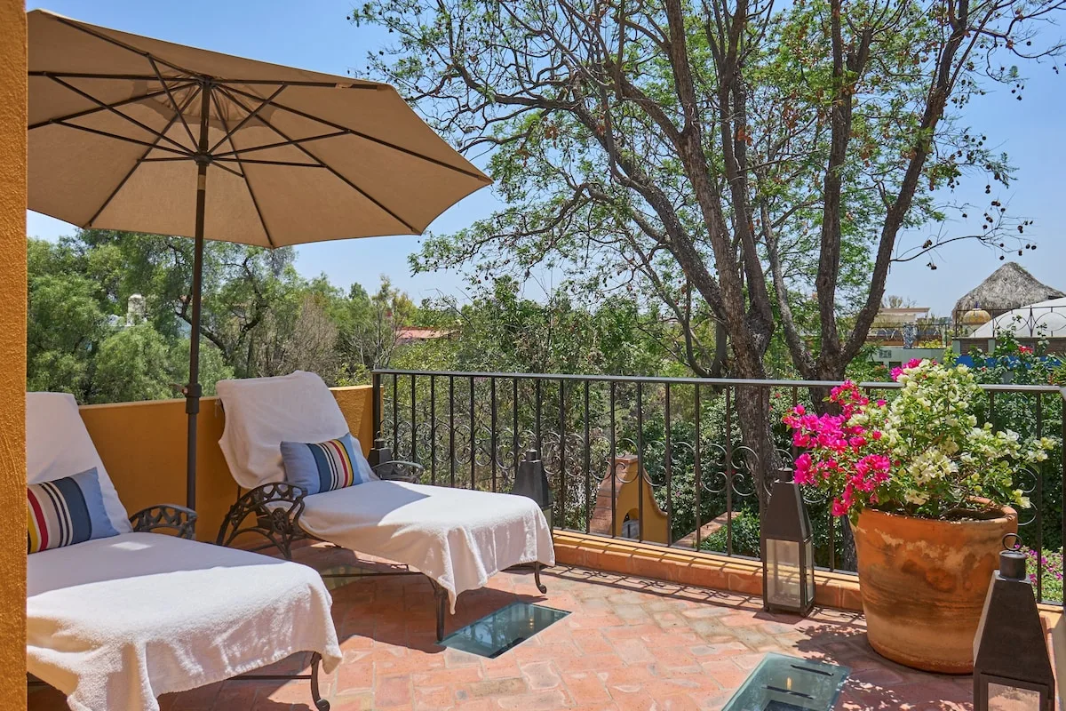 Una terraza al aire libre con asientos acolchados, una sombrilla grande y plantas con flores.