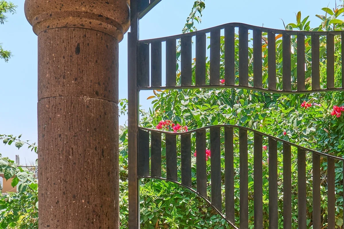 Una puerta de jardín con barrotes oscuros se encuentra entre plantas y flores.