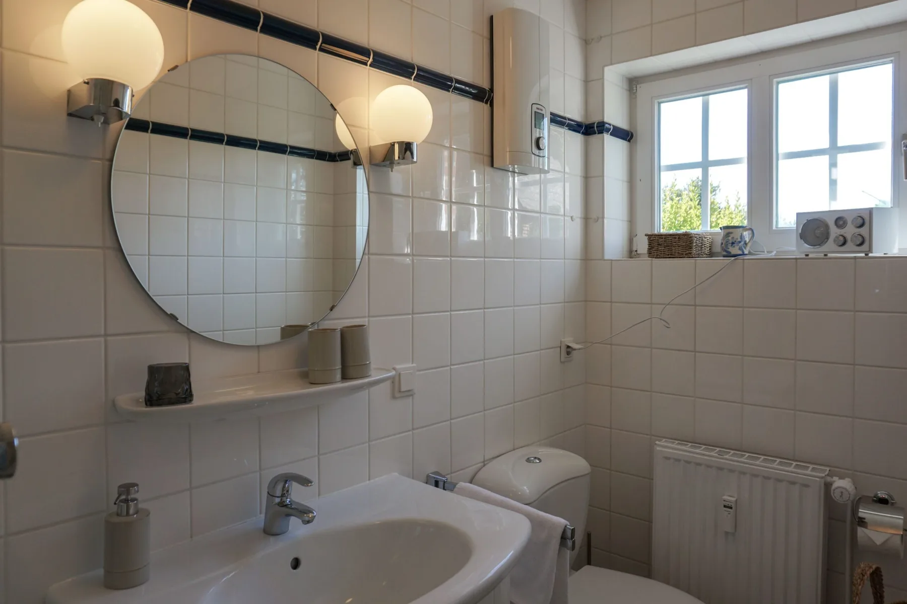 Un baño alicatado con lavabo, espejo redondo, inodoro y una ventana luminosa.