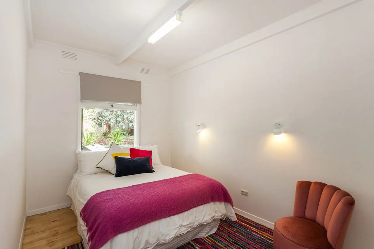 Un dormitorio con una cama, ropa de cama colorida y una pequeña silla tapizada.
