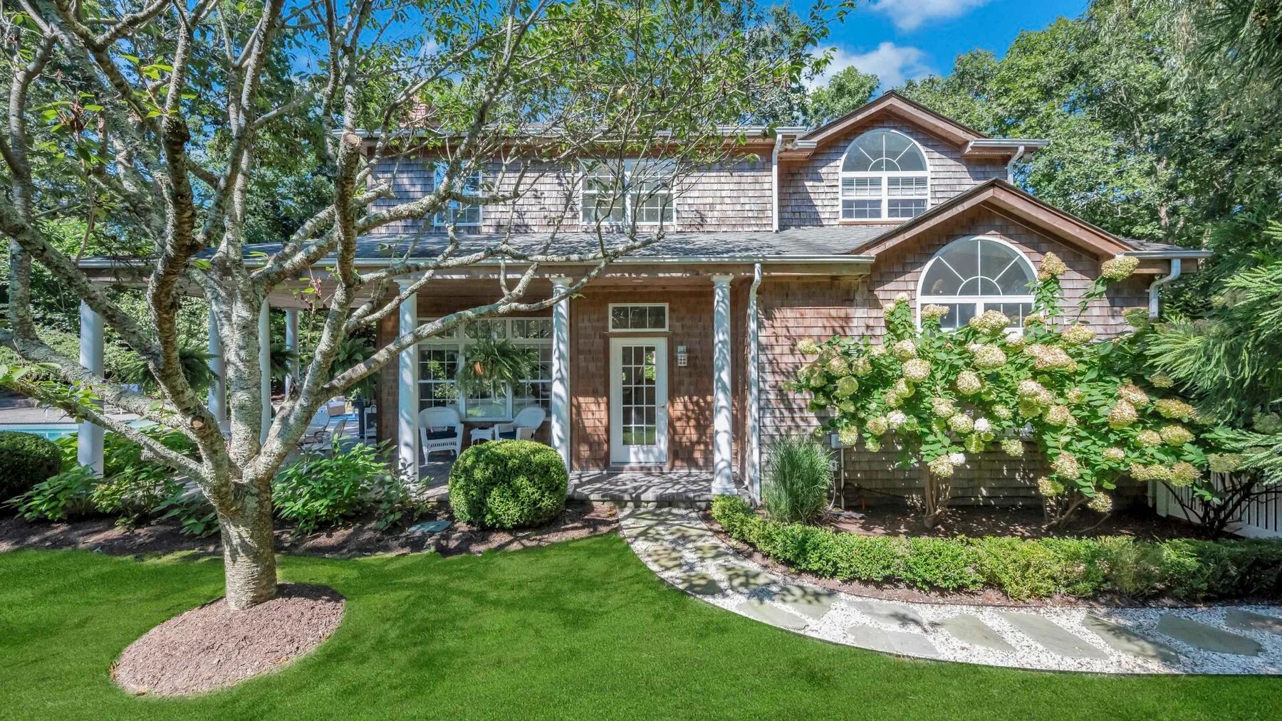 Una casa de dos plantas se alza tras un césped, rodeada de árboles y arbustos floridos.