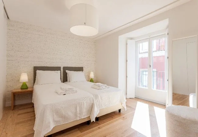 A bright bedroom with two beds beside tall windows.