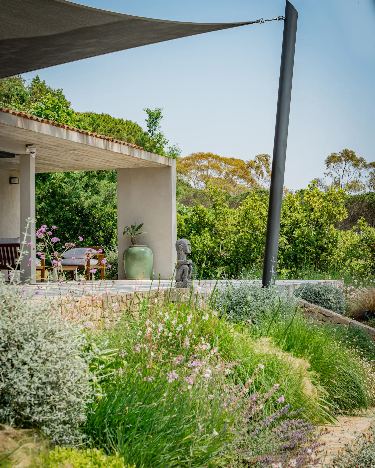 Un patio exterior con sombra da a un jardín frondoso con plantas en flor.