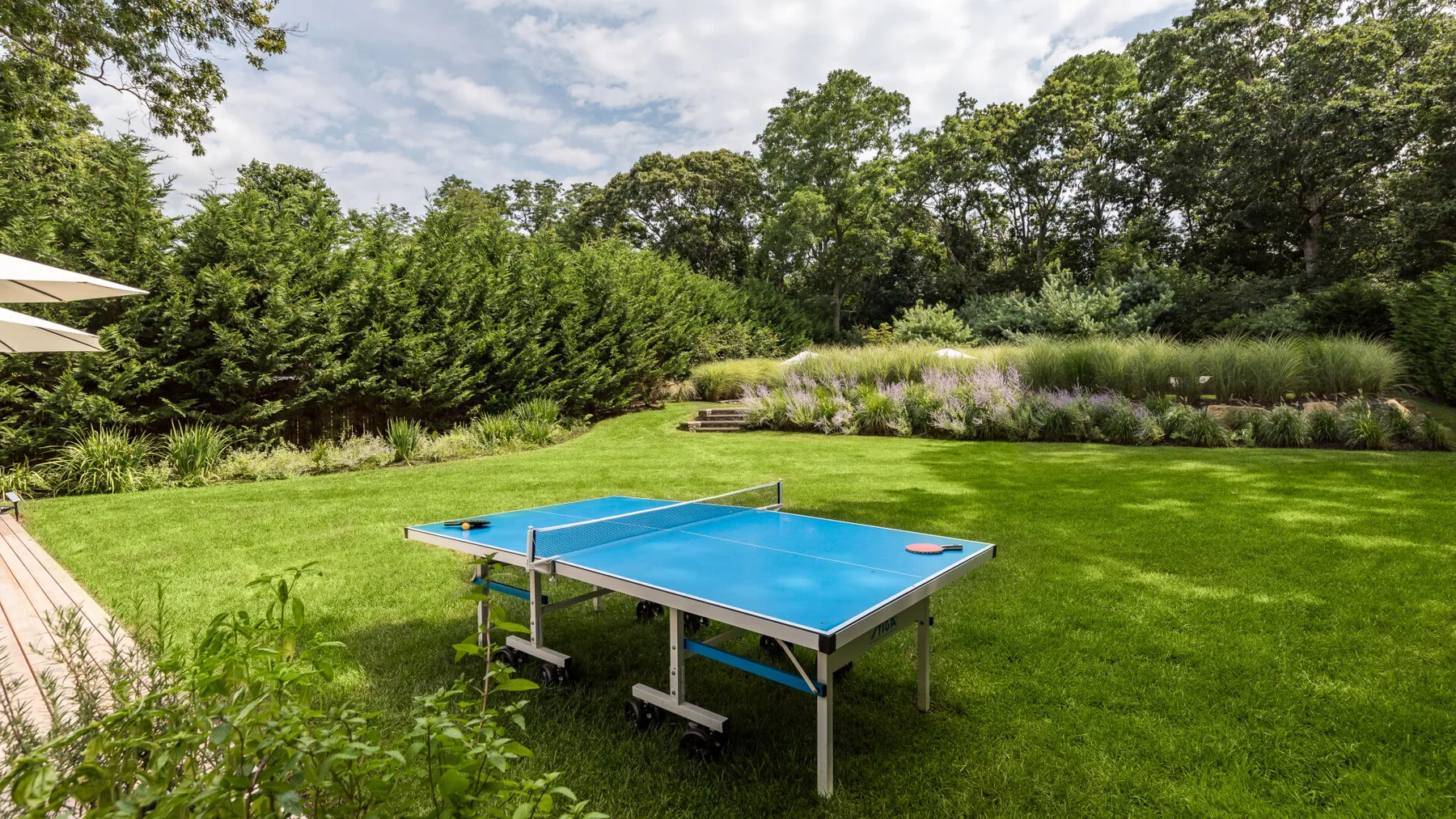 Una mesa de ping-pong azul está sobre el césped, rodeada de árboles y parterres.