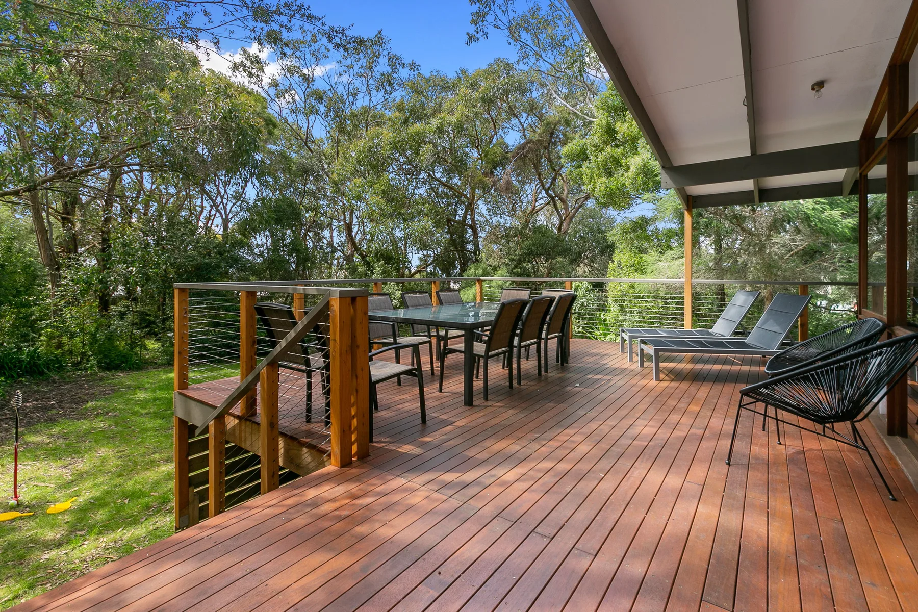 Una terraza exterior de madera tiene una mesa de comedor y asientos entre árboles frondosos.
