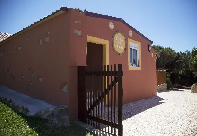 A terracotta-colored building with a small window and a wooden gate beside a gravel yard.