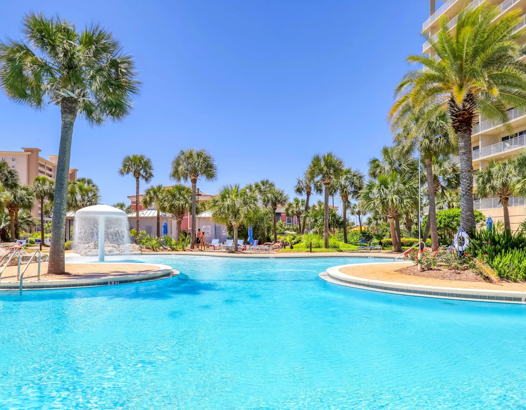 Zona común: Una piscina rodea una fuente, con palmeras y edificios alrededor.