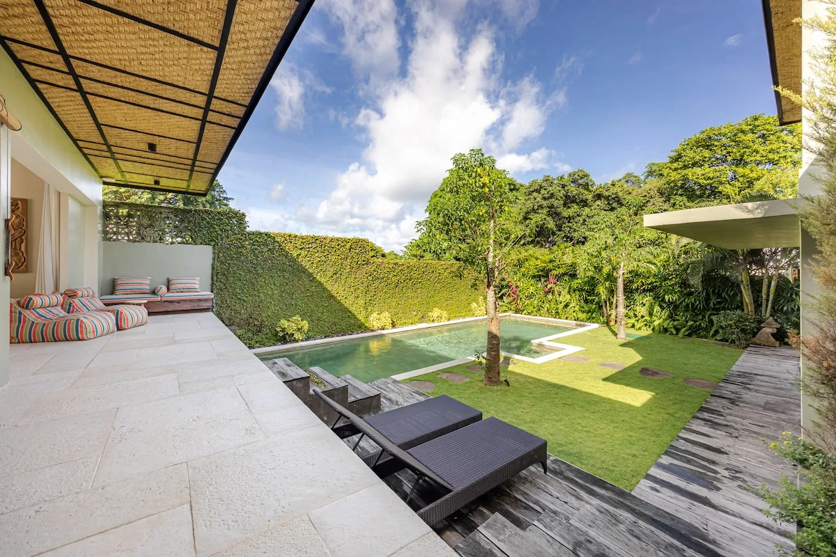 An outdoor patio overlooks a pool, lawn, and leafy garden.