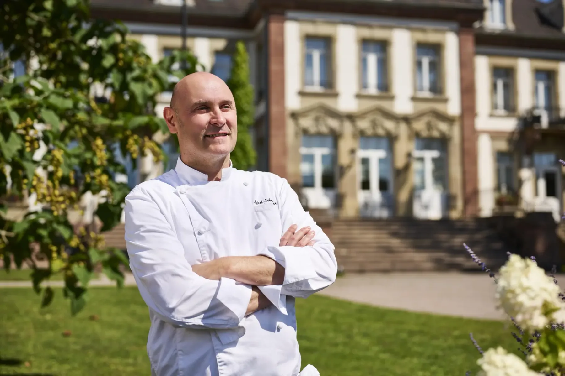 Representative hotel space: A chef stands with folded arms in a garden outside a large building.