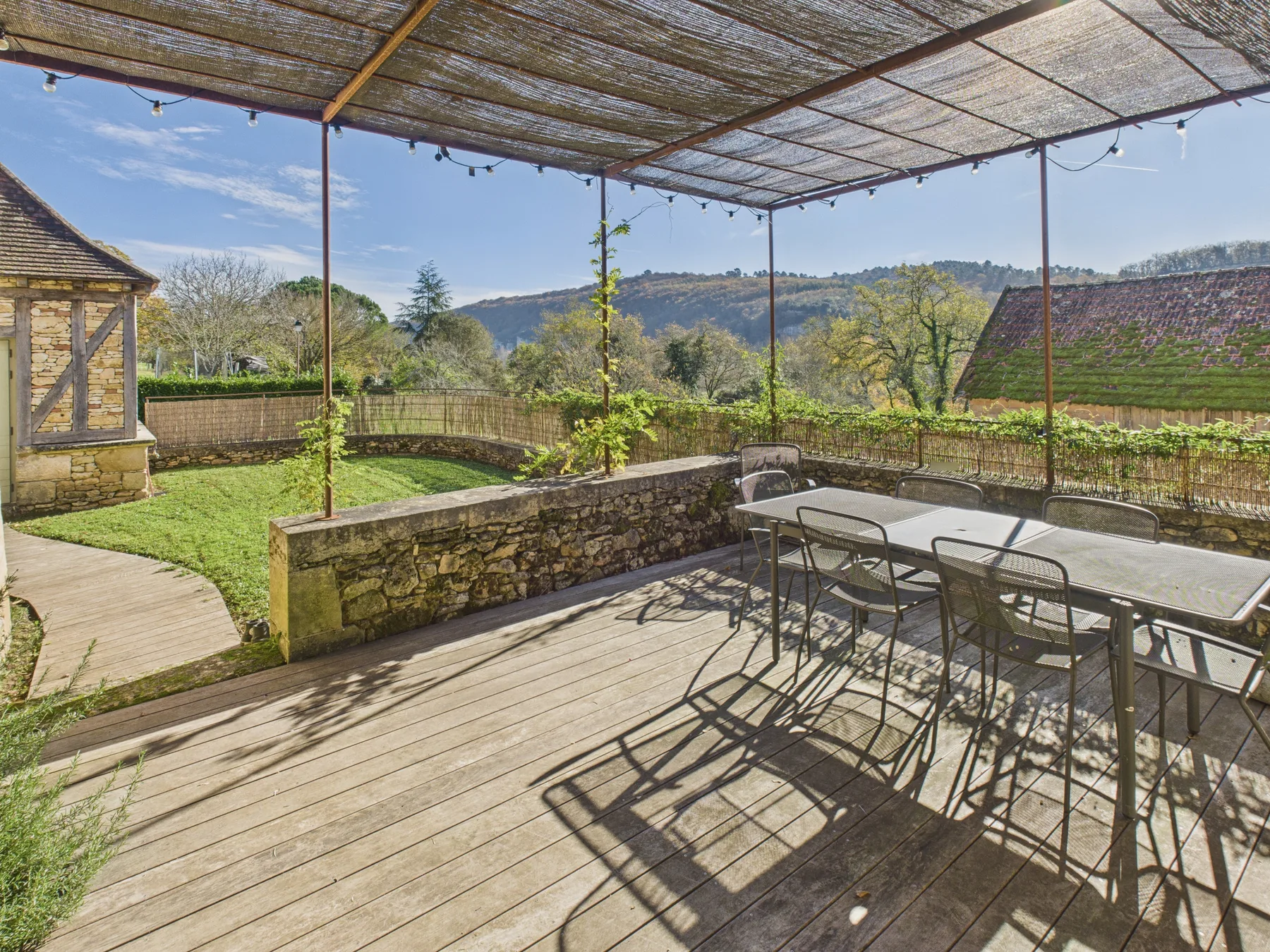 A covered outdoor terrace has a dining table beside a garden and distant hills.