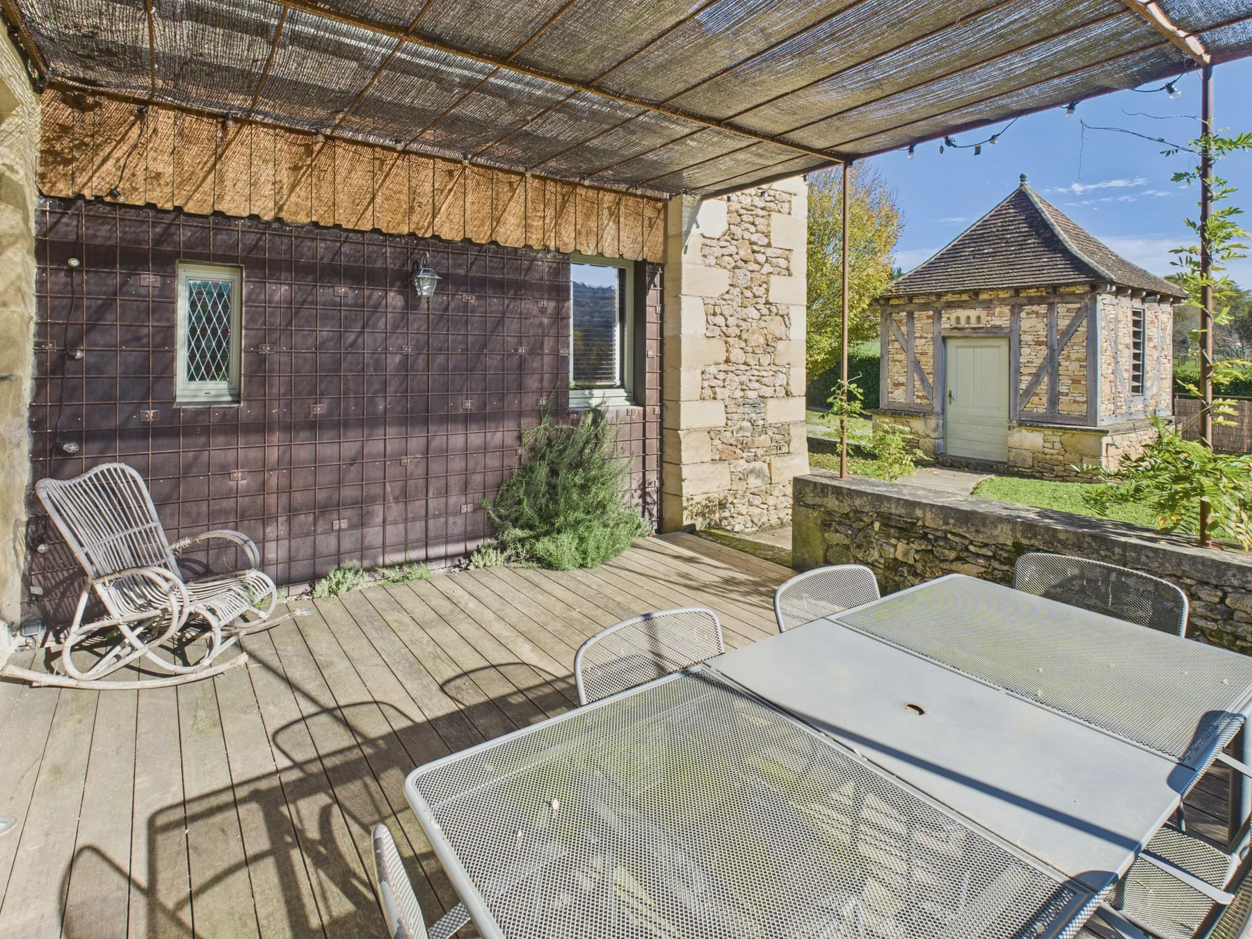 An outdoor patio with a shaded dining table and seating beside a stone building.