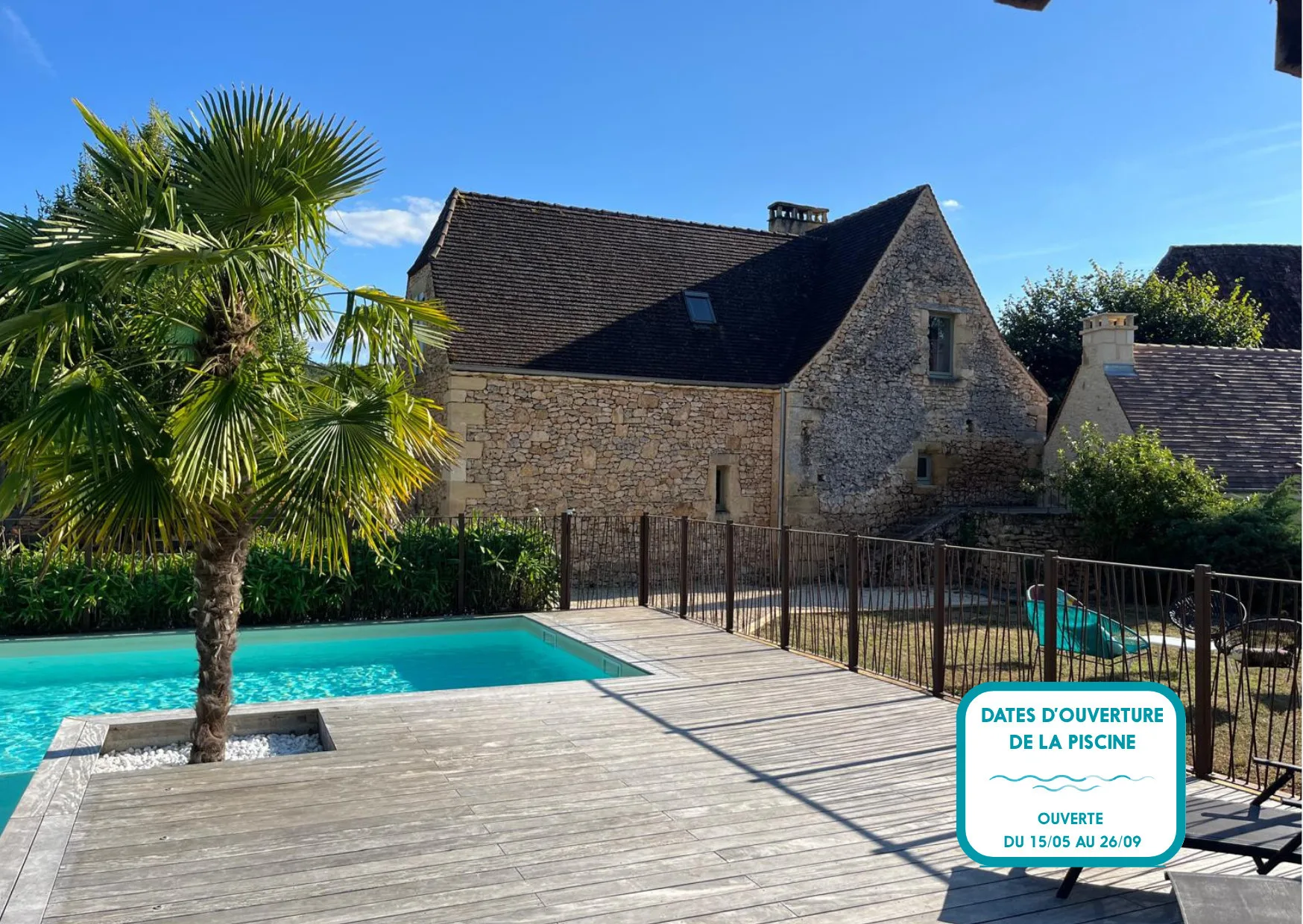 An outdoor pool and wooden deck sit beside a stone house and palm tree.