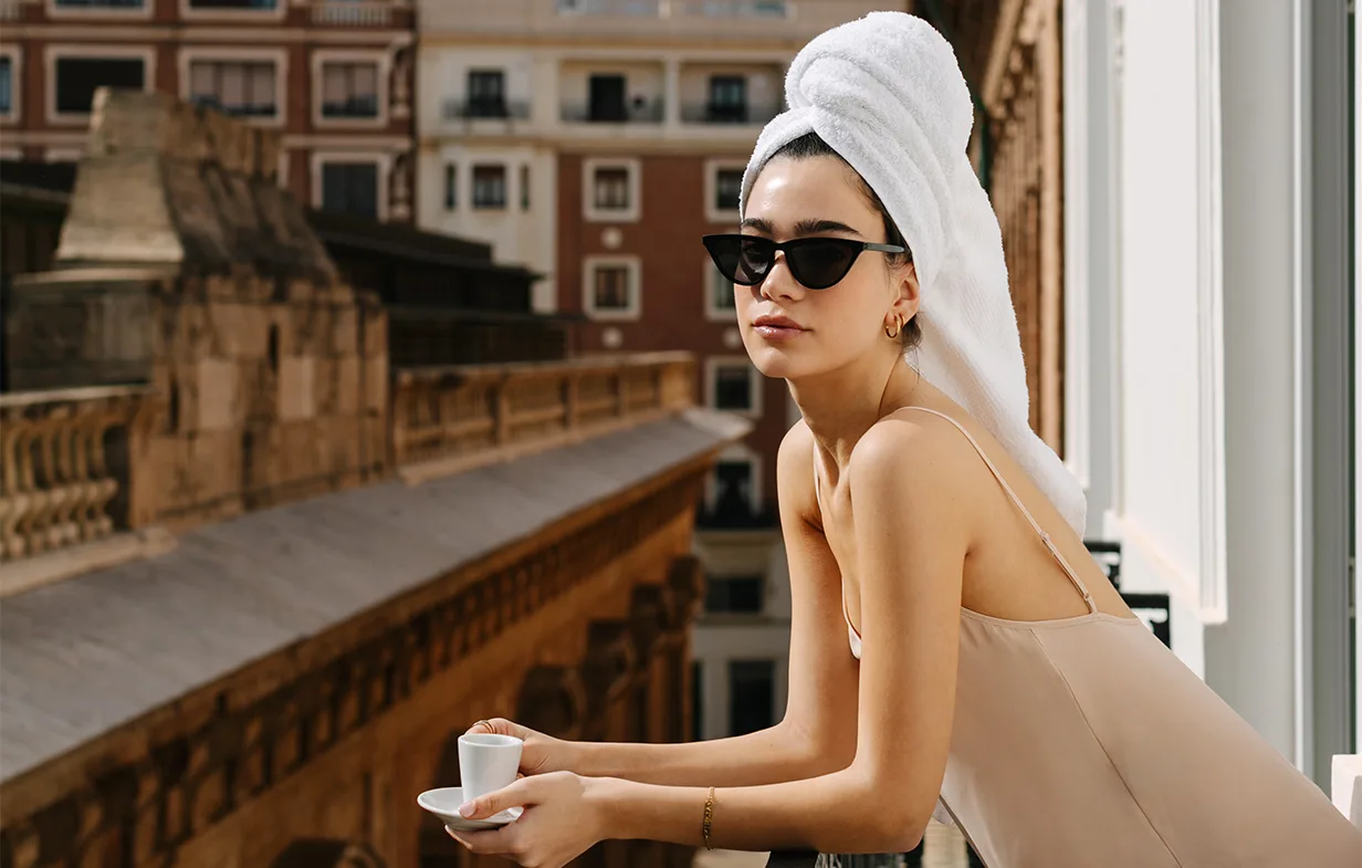 Representative hotel space: A woman wearing sunglasses and a towel sits by a balcony railing with a cup.