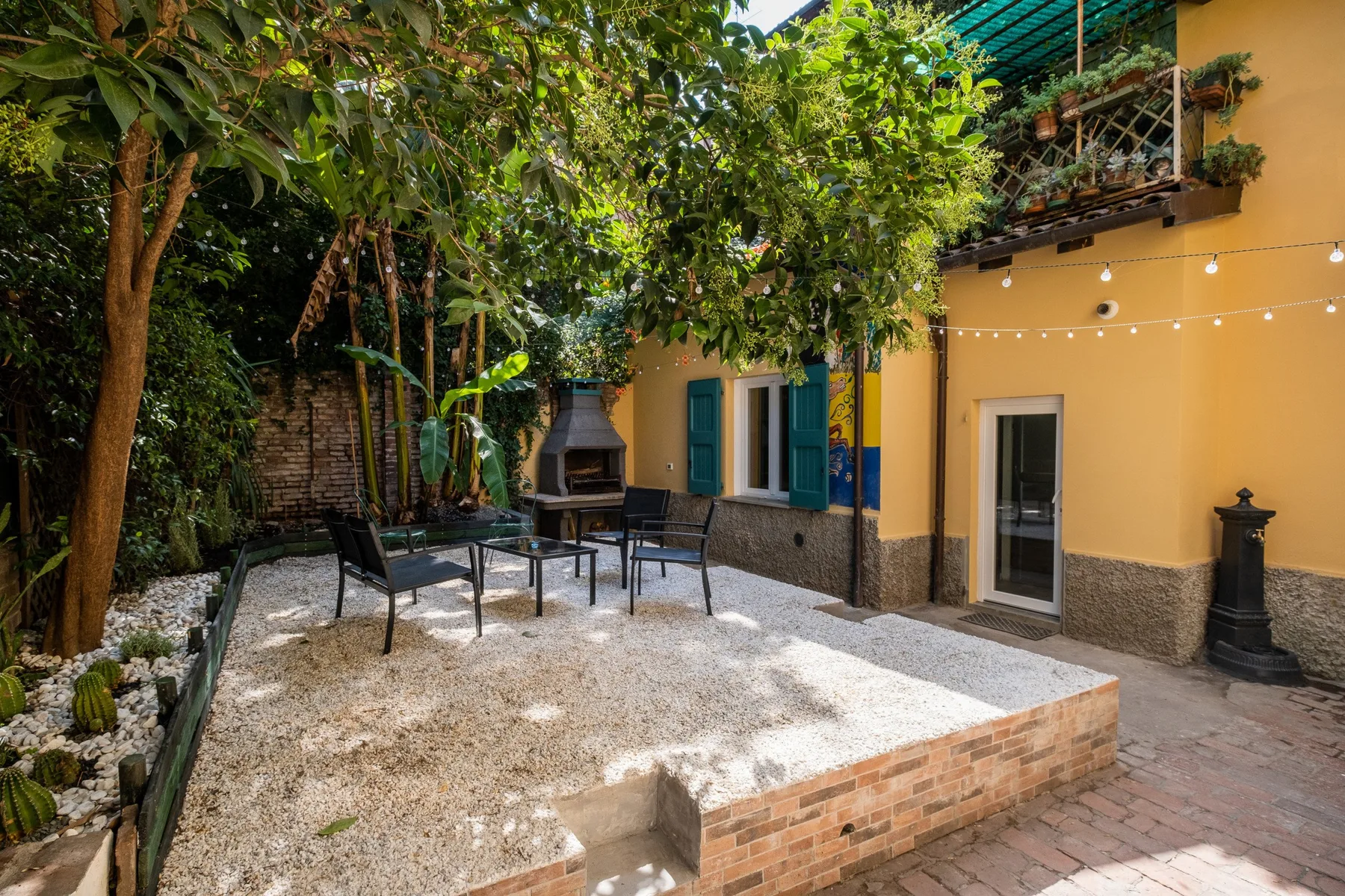 Patio exterior con asientos bajo árboles frondosos junto a un edificio amarillo.