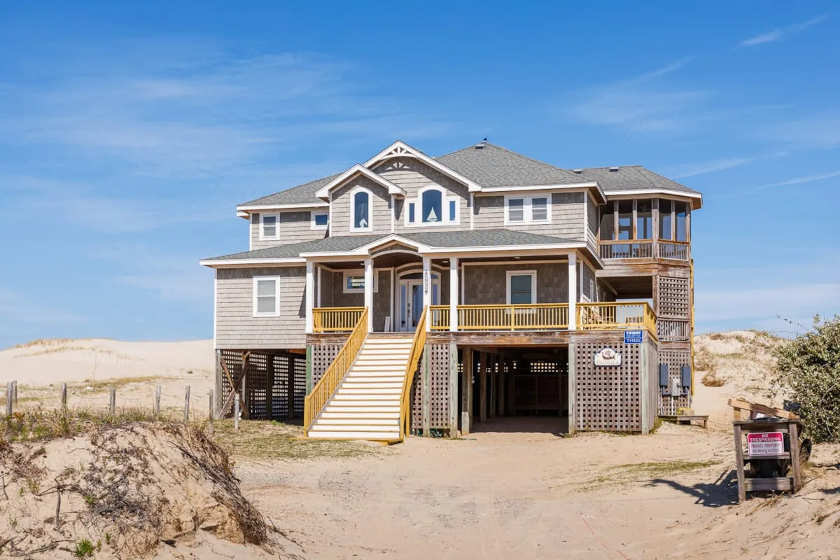 A large beach house stands among sandy dunes beneath a clear blue sky.