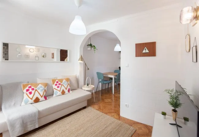 Una sala luminosa con un sofá, cojines estampados y un arco que da a un comedor.
