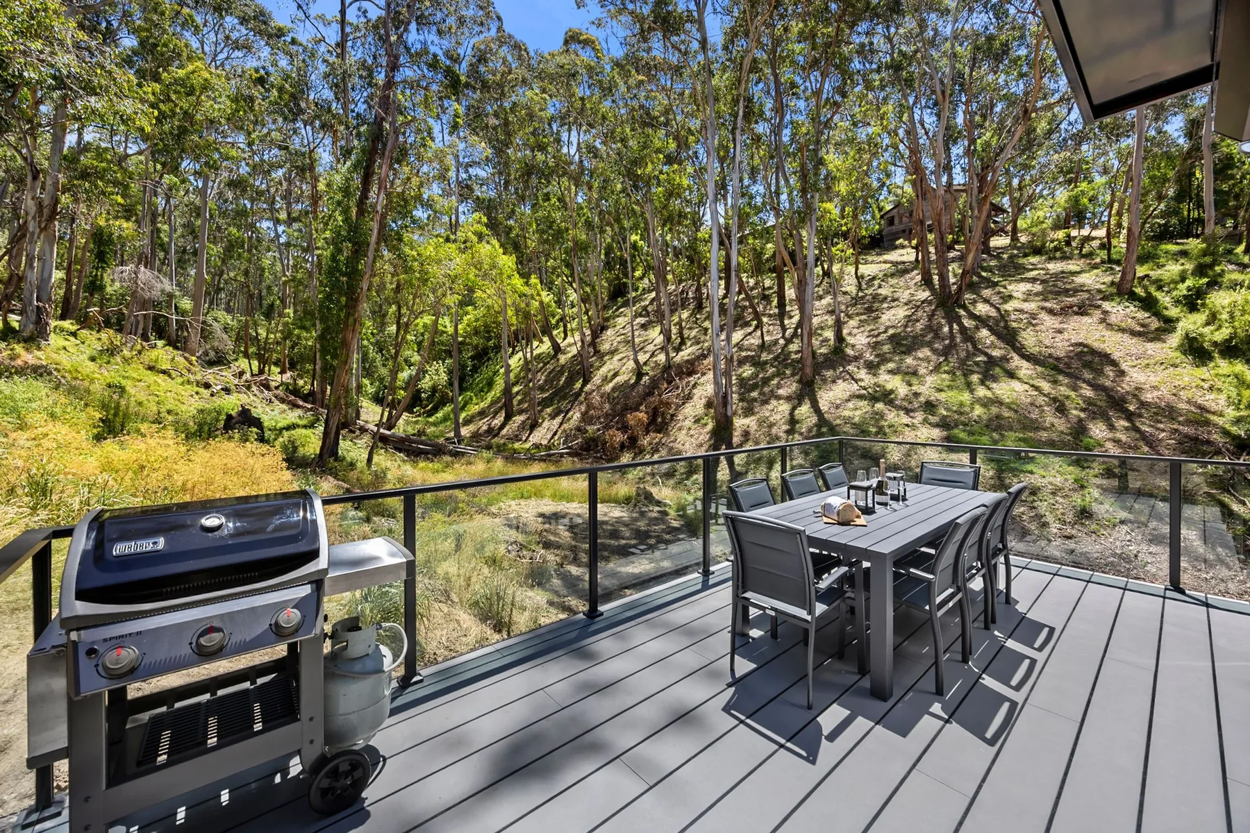 Una terraza exterior tiene una mesa de comedor y una parrilla junto a una ladera arbolada.