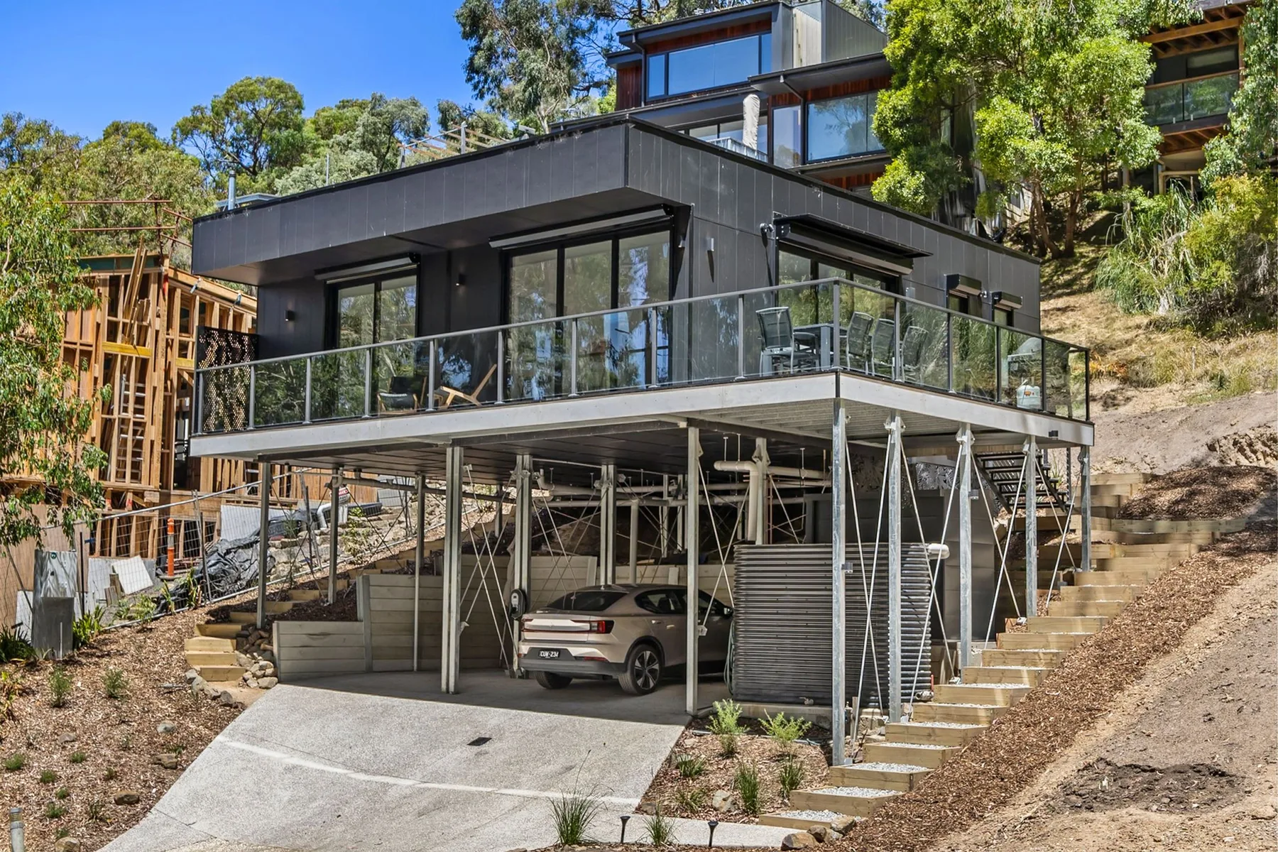 Una casa moderna está en una ladera, con un balcón de vidrio sobre un garaje abierto.