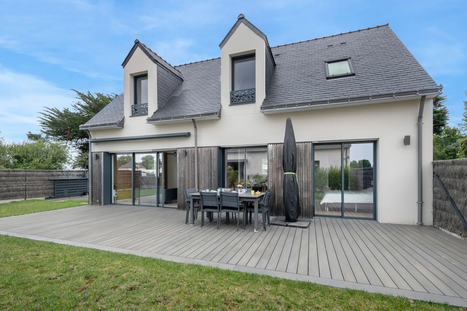 A modern house opens onto a broad deck with outdoor dining seating.