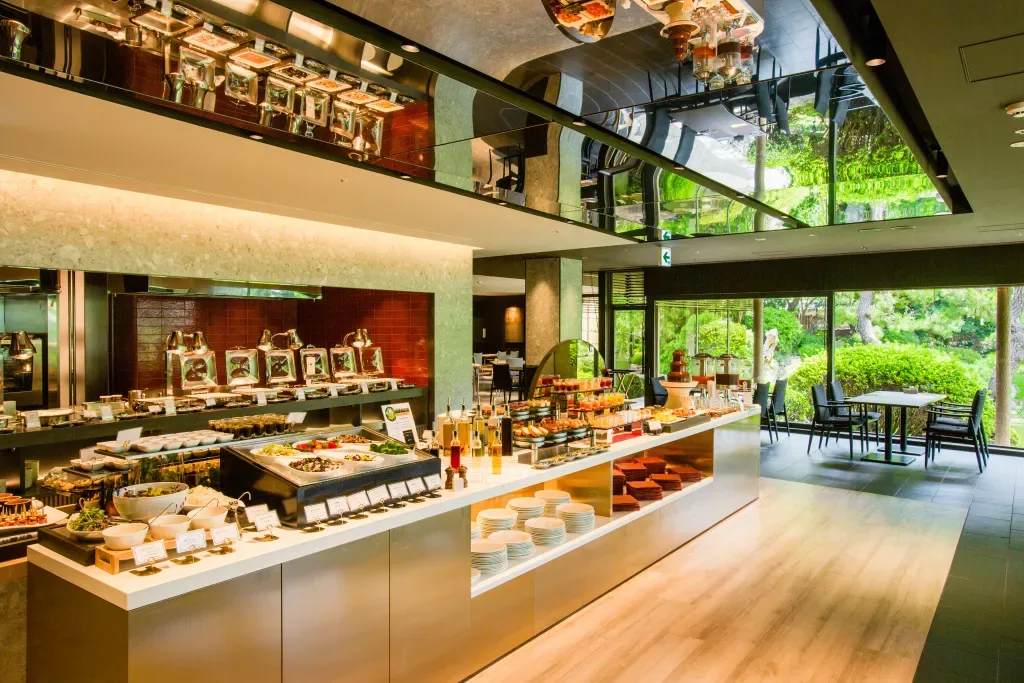 Espacio representativo del hotel: Área de bufé con exhibidores de comida, un mostrador y mesas junto a grandes ventanas.