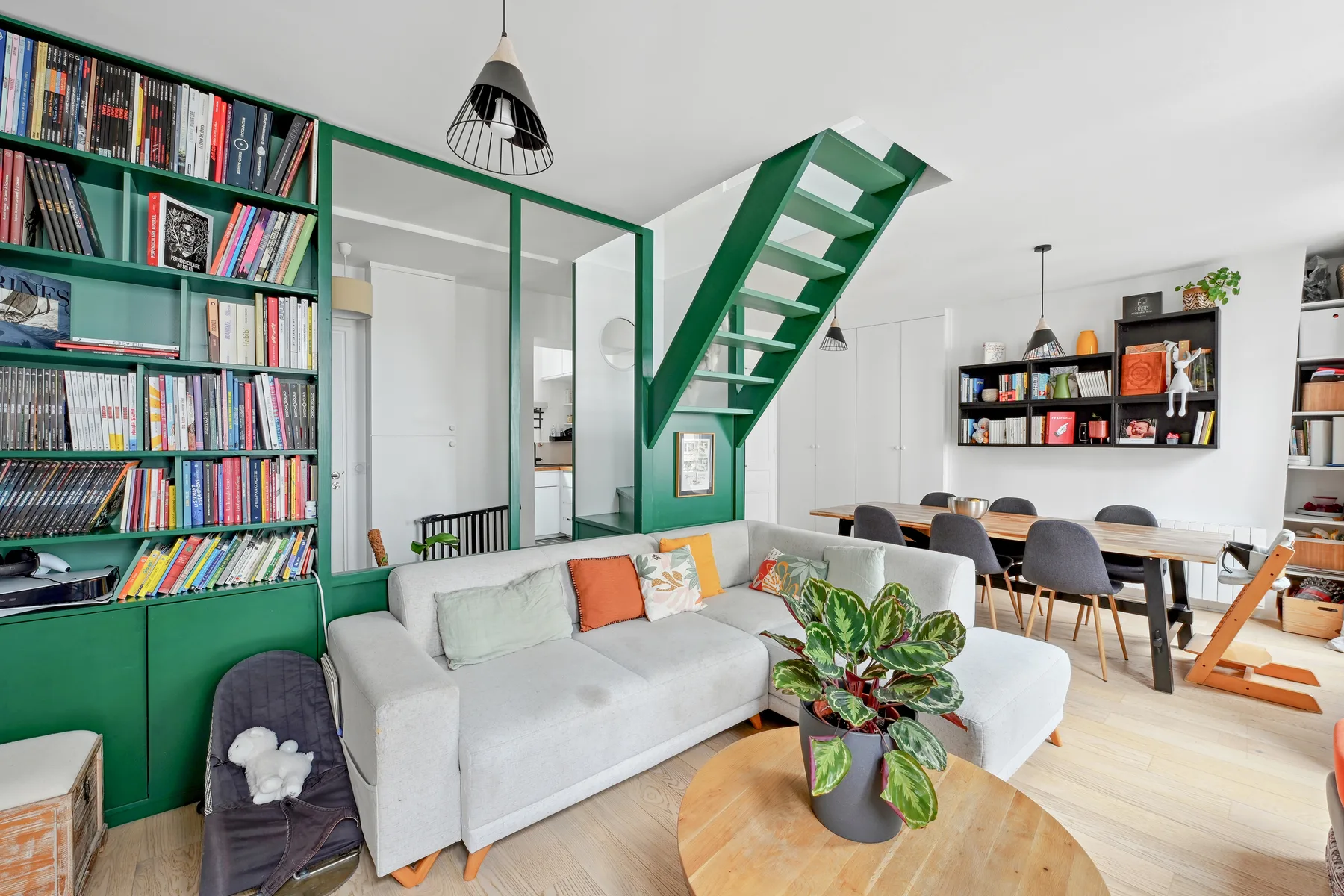 A bright living room with a gray sofa, green shelving, and an open staircase.