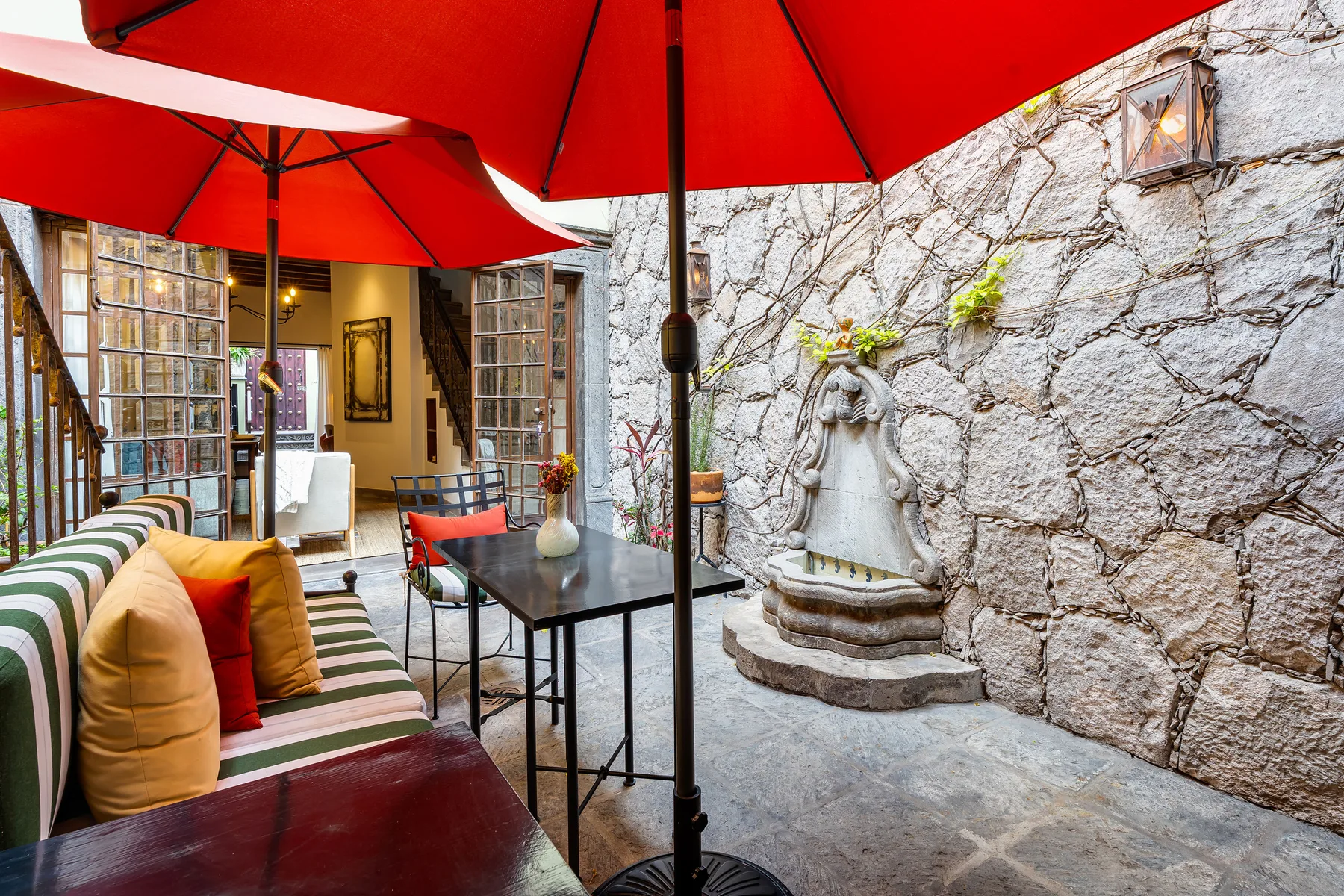 A courtyard seating area has striped cushions, red umbrellas, and a stone fountain.