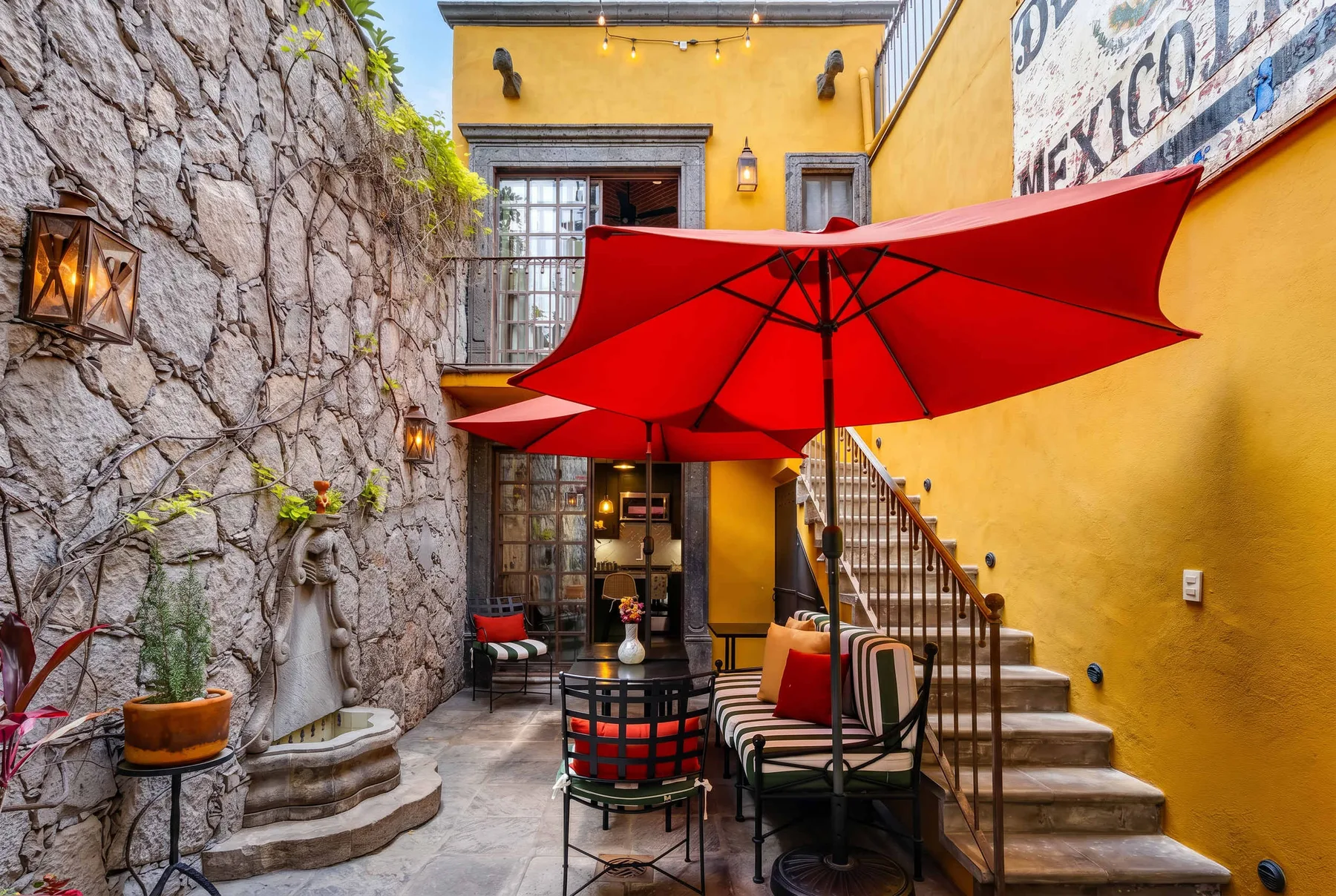 An outdoor courtyard with red umbrellas, seating, and a staircase beside yellow walls.