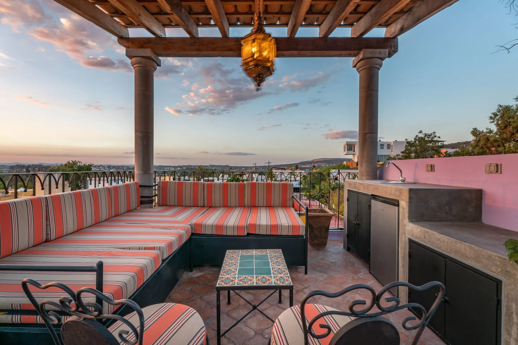 An outdoor terrace has striped seating, a low table, and a built-in counter beneath a wooden pergola.
