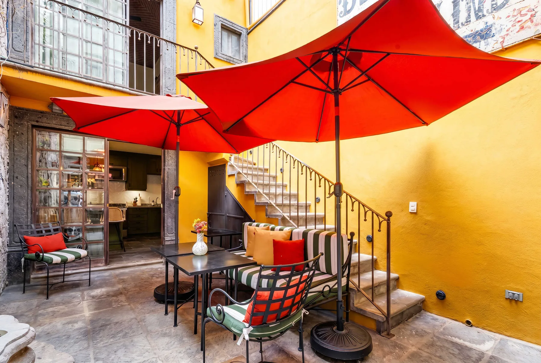 A courtyard seating area with red umbrellas and a staircase beside a yellow building.