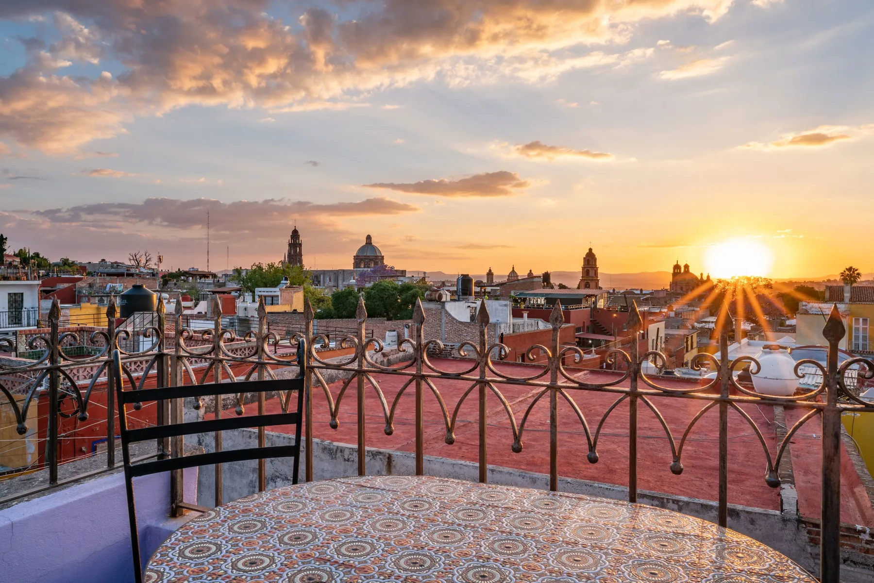 A rooftop terrace overlooks a city skyline at sunset, with patterned tiles in the foreground.