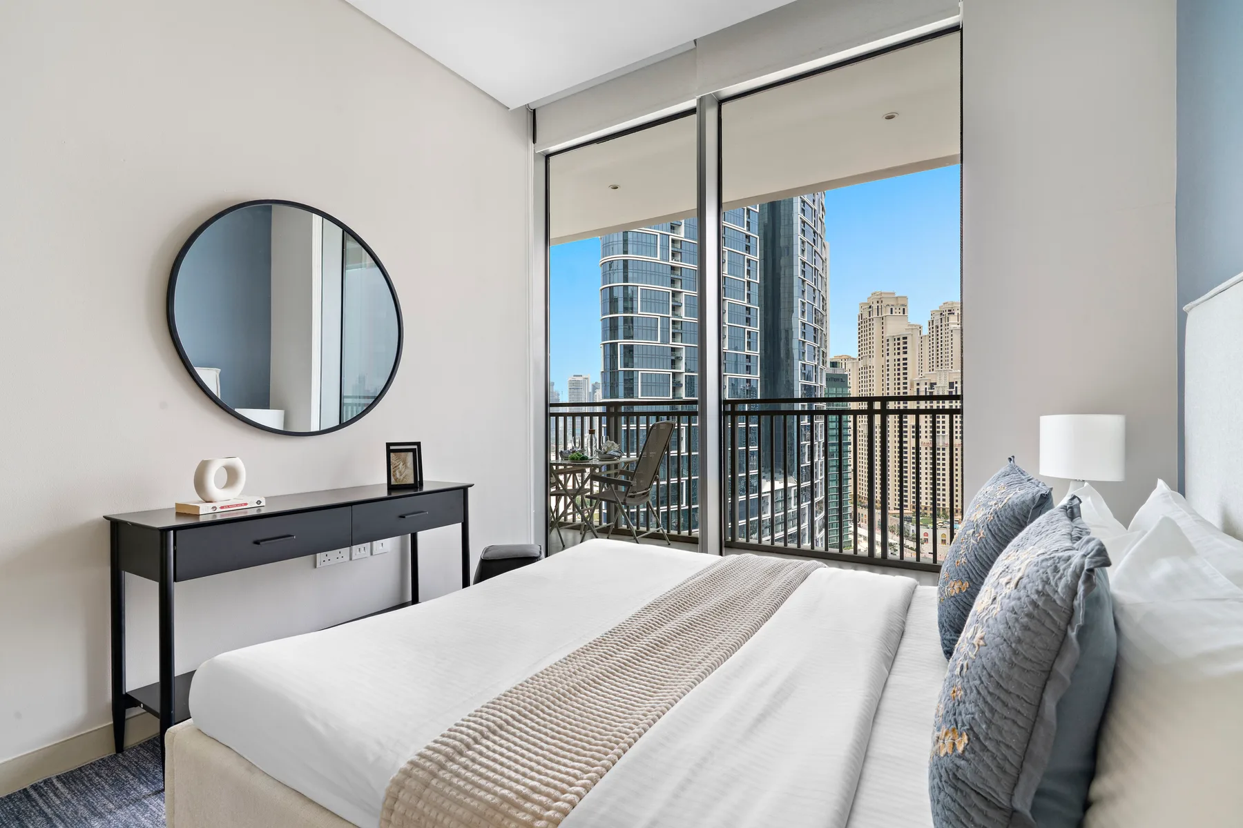Un dormitorio luminoso con una cama junto a ventanales con vistas a edificios de la ciudad.