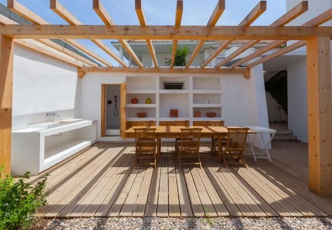 Un comedor al aire libre bajo una pérgola de madera, con una mesa larga y asientos.