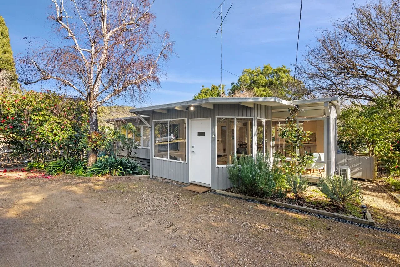 A small house sits beside a dirt yard, surrounded by trees and garden plants.