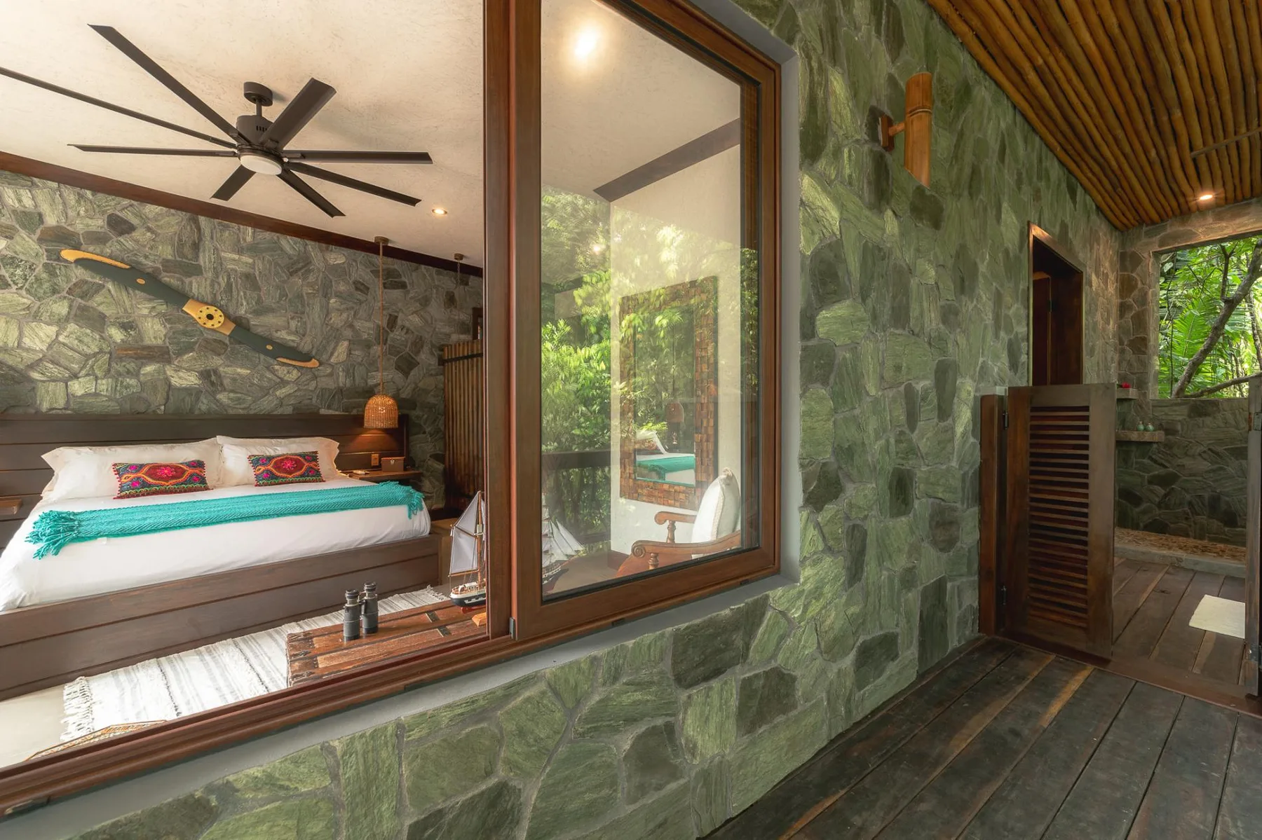A bedroom with a bed is visible through a large window beside a stone wall.