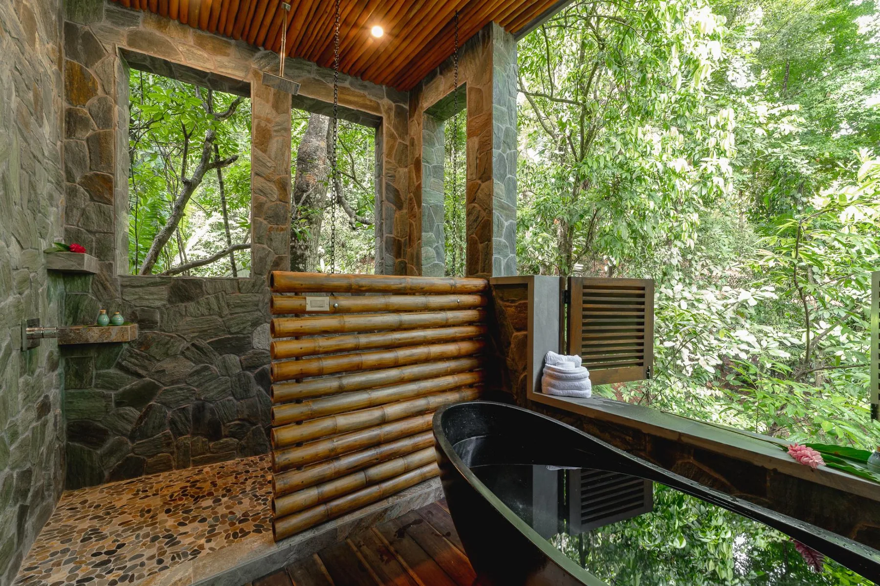 A bathroom with a black soaking tub, stone walls, and leafy views through large windows.