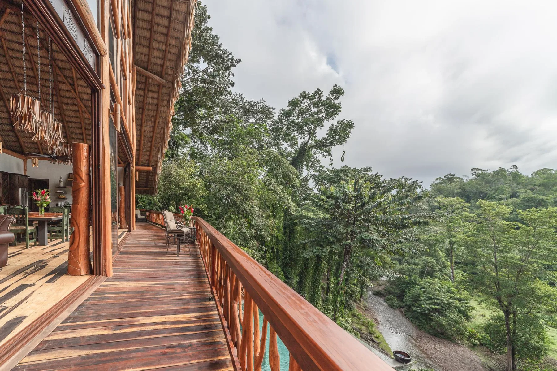 A wooden balcony overlooks dense green trees beneath a cloudy sky.