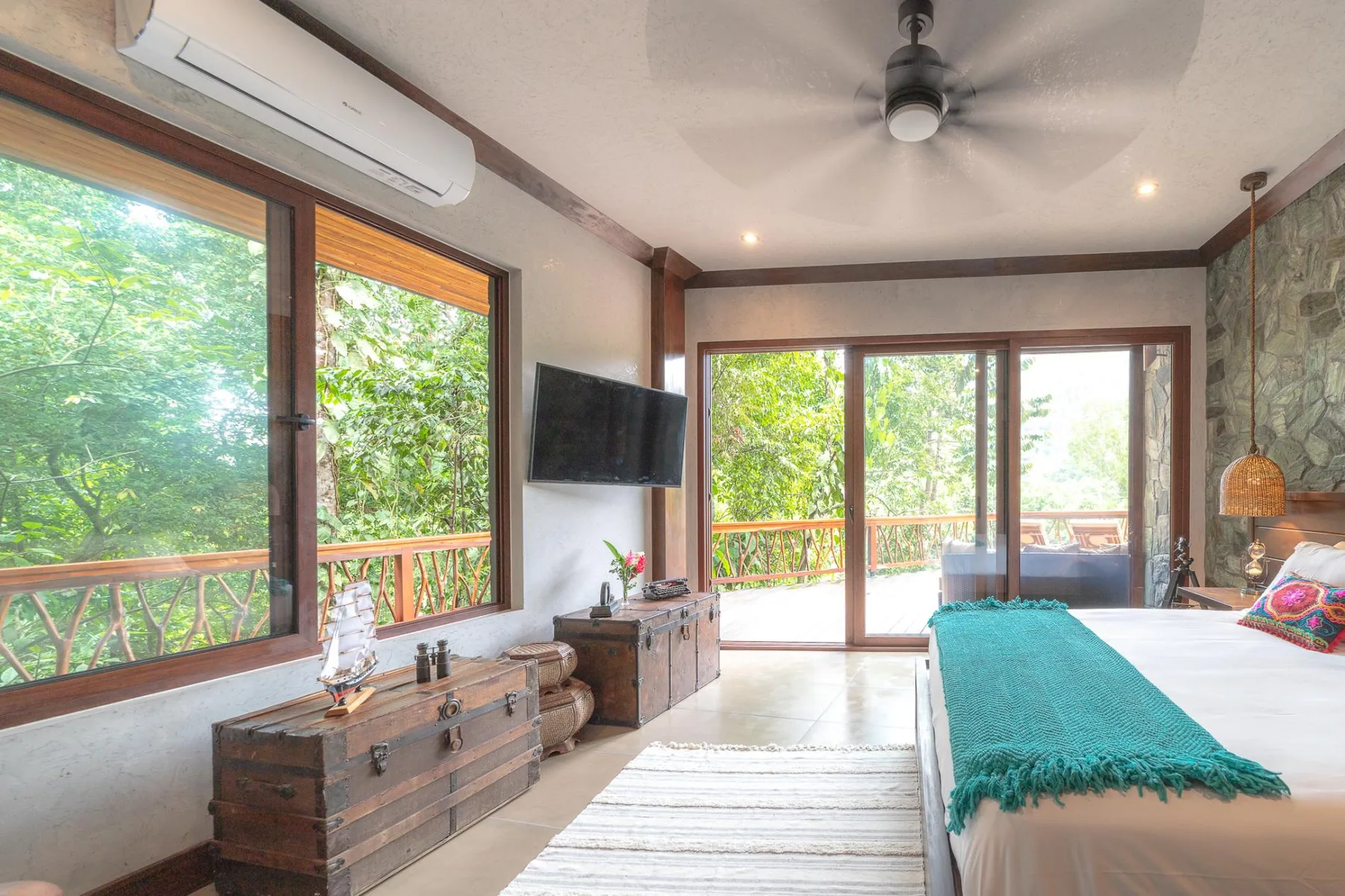 A bedroom with a bed beside large windows overlooking leafy greenery.