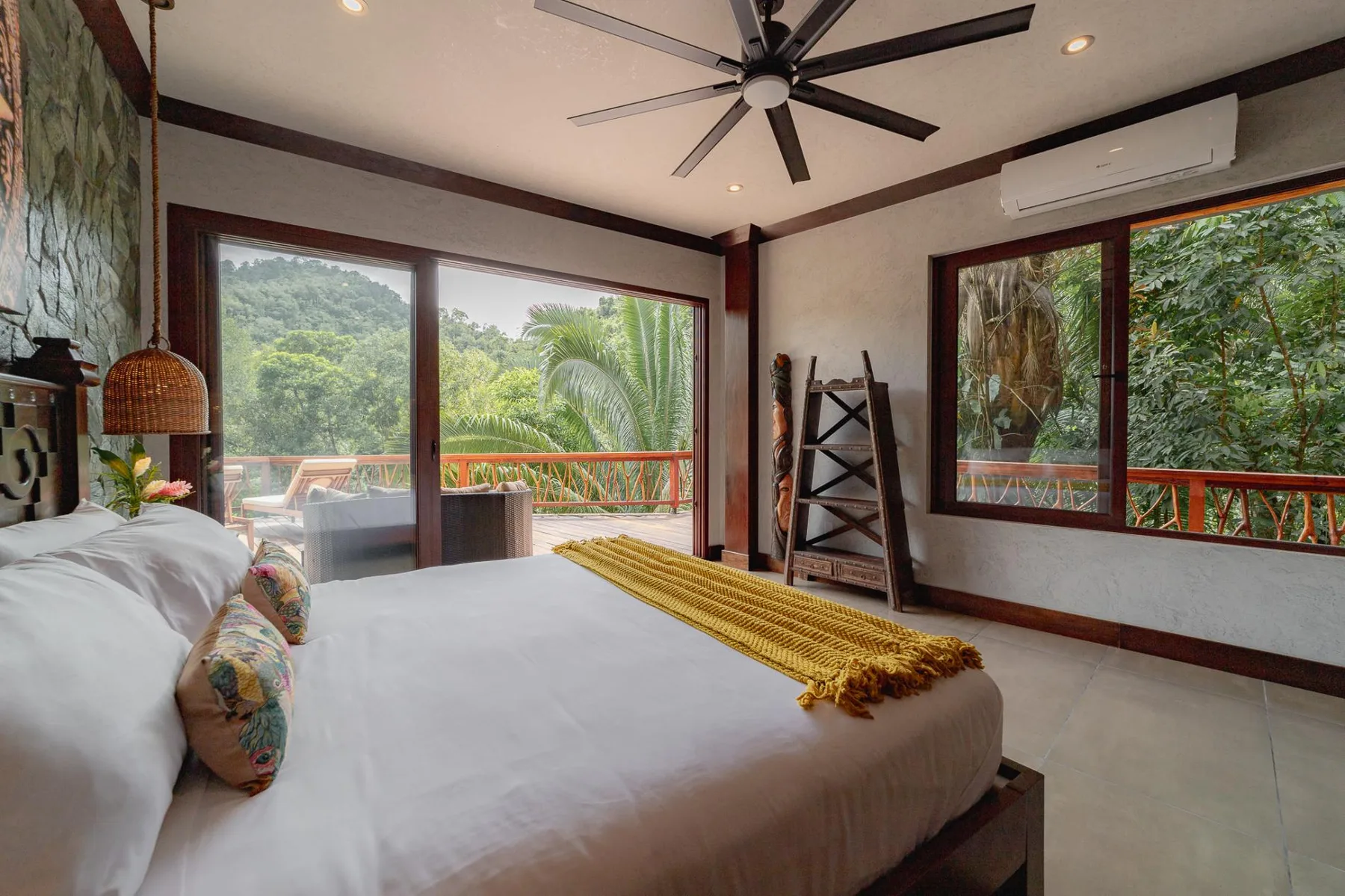 A bedroom with a bed beside wide windows overlooking lush greenery.