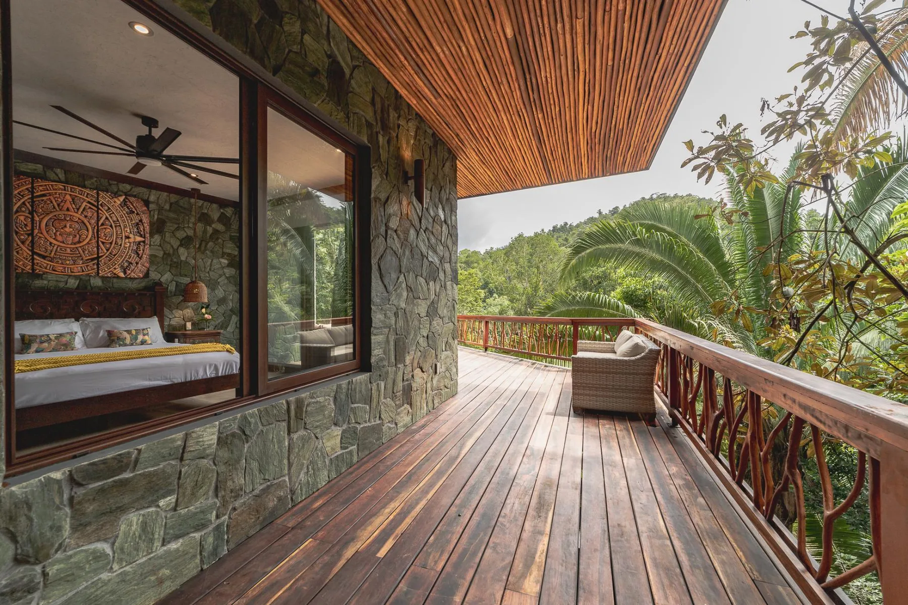 A wooden balcony beside a bedroom overlooks lush green trees.