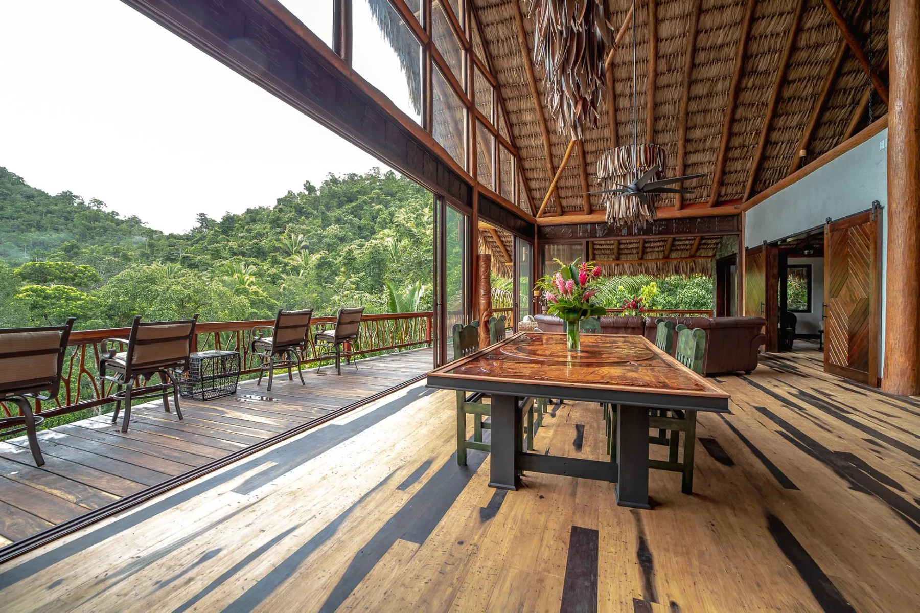 A covered wooden terrace has a long table and seating overlooking a green hillside.