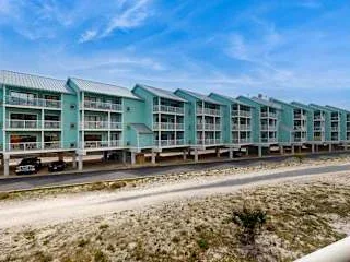 Un largo edificio de apartamentos color turquesa con balcones se alza junto a una playa arenosa.