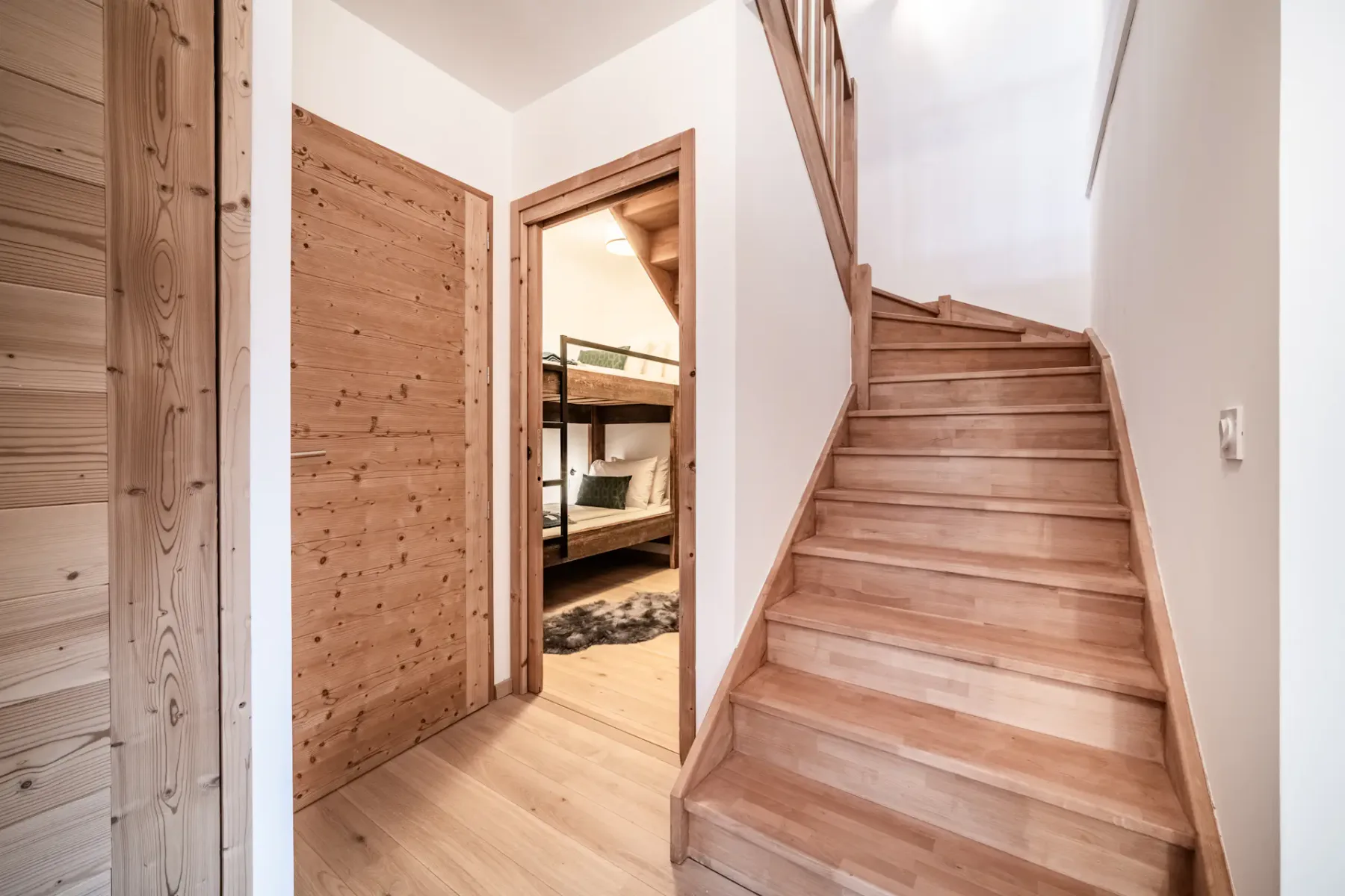 Una escalera de madera sube junto a una puerta abierta que da a una habitación con literas.