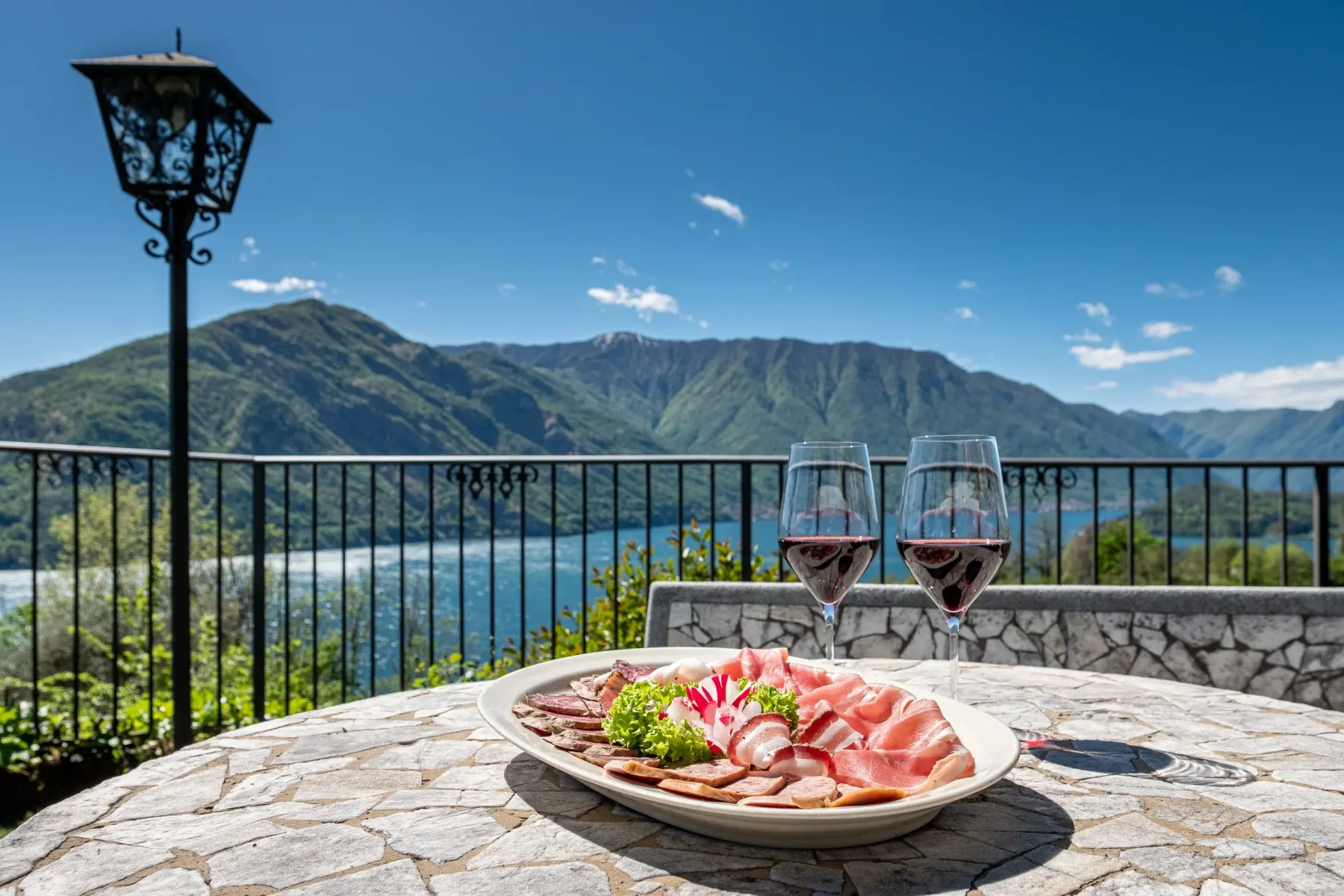 Representative hotel space: A plate of sliced food and two glasses sits on a stone terrace overlooking a lake and mountains.