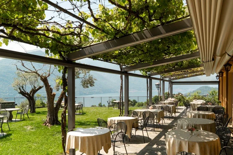 Representative hotel space: Outdoor dining area with tables beneath a leafy pergola beside the lake.