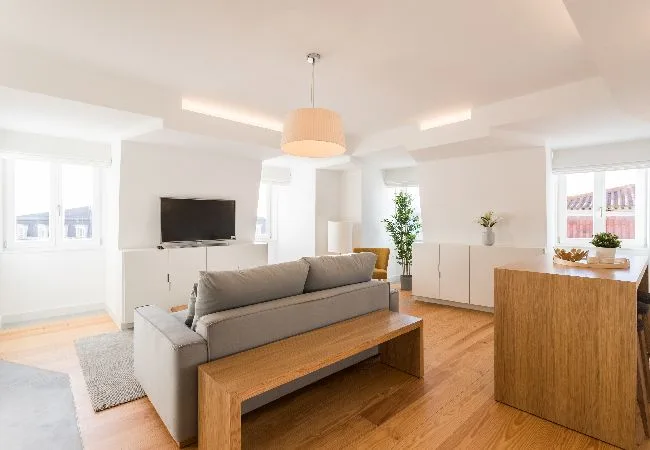 Una sala luminosa con un sofá gris, una mesa de centro de madera y un televisor.