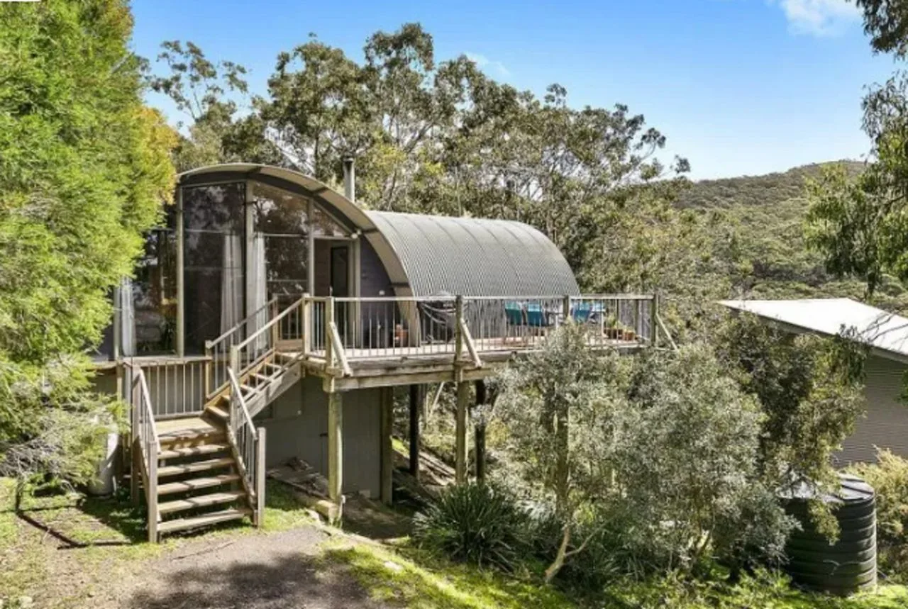 Una cabaña elevada con techo curvo y una terraza exterior entre árboles.