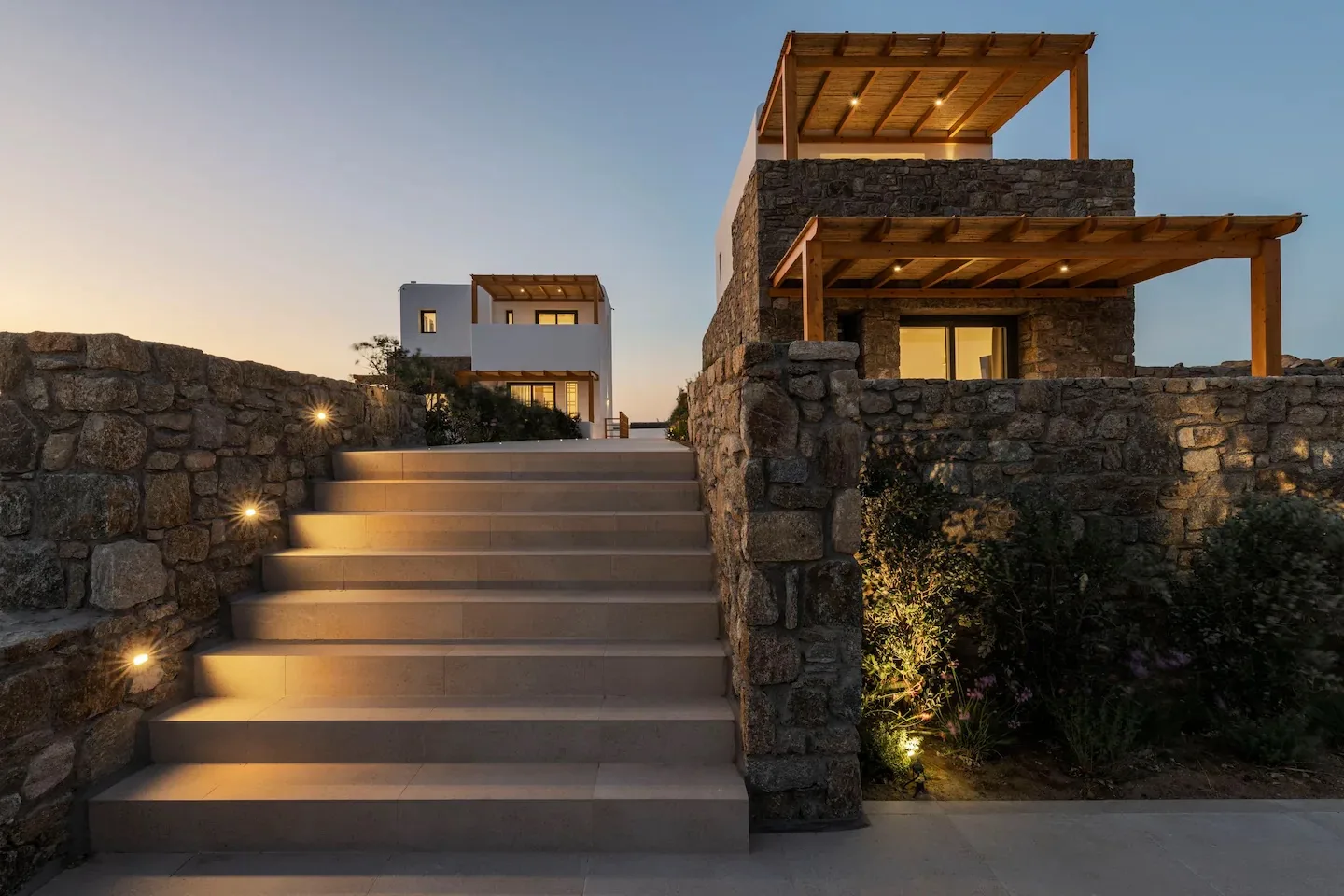 Unas escaleras de piedra conducen a una casa moderna con zonas exteriores iluminadas al anochecer.