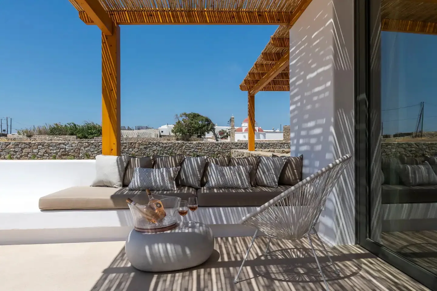 Un patio al aire libre tiene asientos acolchados bajo una pérgola de madera.